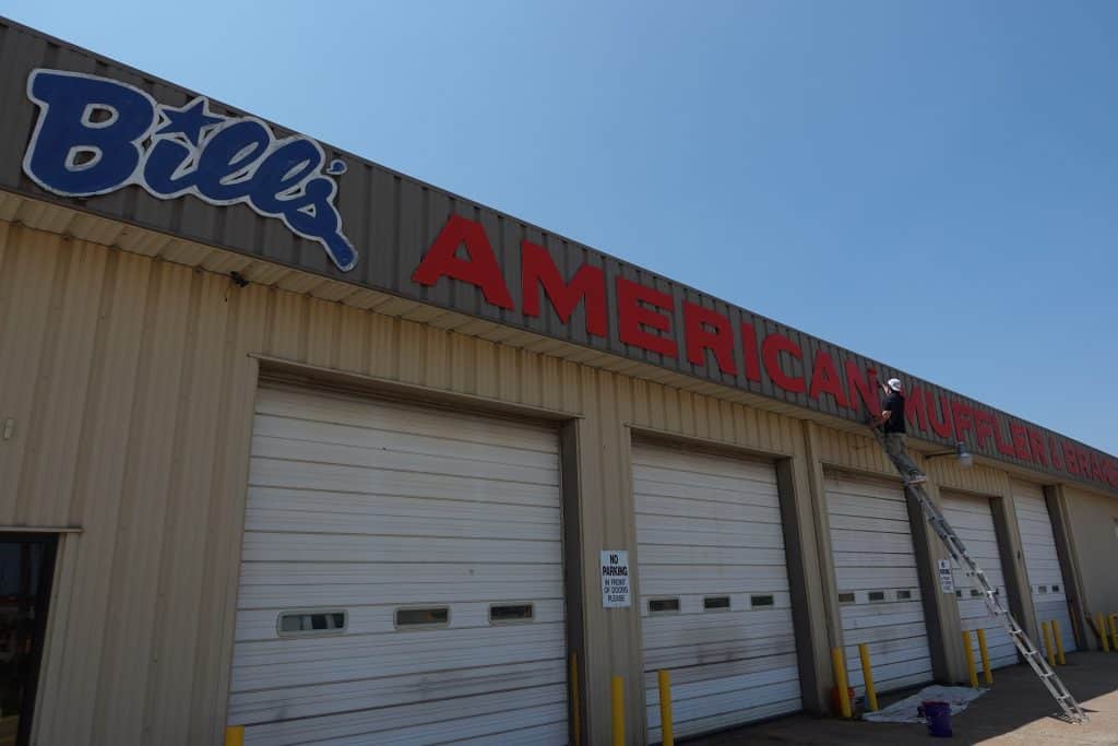 A professional painter in Wichita, KS, working on a large commercial building sign to refresh its appearance. The bold red and blue lettering contrasts with the neutral exterior, creating a vibrant, professional look.